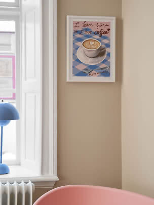 Gerahmtes Kaffeeposter mit Latte Art und Text an beiger Wand neben einem Fenster
