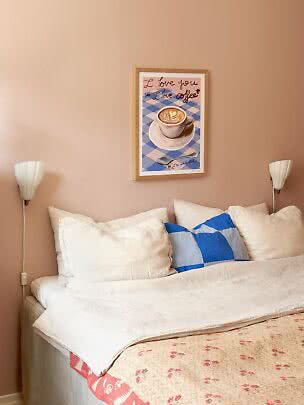 Bett mit weißen Kissen, blauem Zierkissen und Kaffeeposter an rosafarbener Wand