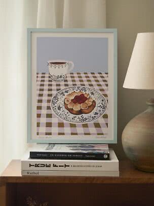 Stampa da cucina incorniciata con tazza e dolce su tovaglia a quadri, su libri accanto a una lampada