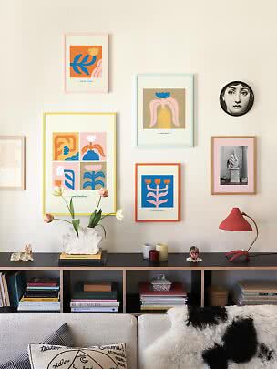 Parete del soggiorno con stampe colorate incorniciate sopra una credenza nera con vaso, tulipani e lampada rossa