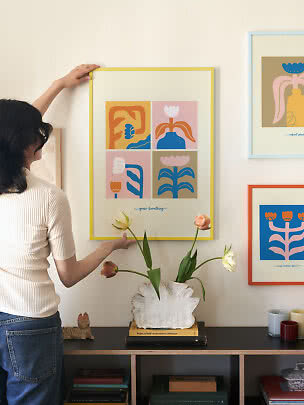 Persoon hangt kleurrijke bloemenposter in gele lijst boven kast met vaas en tulpen