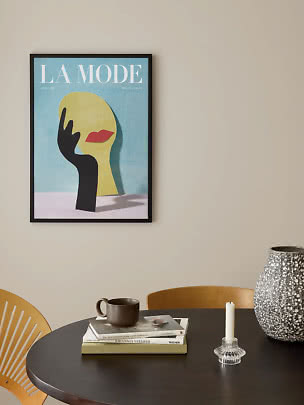Affiche de mode encadrée avec visage abstrait et main noire sur mur beige au-dessus d’une table ronde