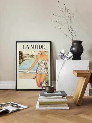 Framed vintage fashion poster leaning against wall with books, cup, black vase and wooden stool