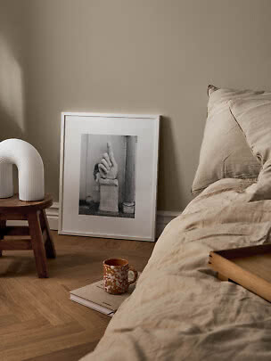 Gemütliches Schlafzimmer mit beiger Bettwäsche, weißer Lampe auf Hocker und gerahmter Kunst am Boden