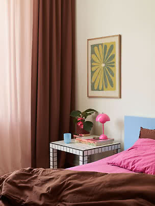 Schlafzimmer mit braunen Vorhängen, pinkem Bett, Fliesentisch, pinker Lampe und Blumenbild