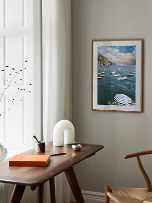 Framed seaside print with boats above a wooden desk in a bright, minimalist room
