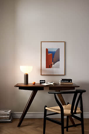 Dark wooden desk with glowing table lamp, books, notebook and black chair beneath framed wall art.