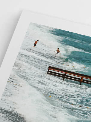 Framed beach photo with two surfers in the waves and a partially submerged bench