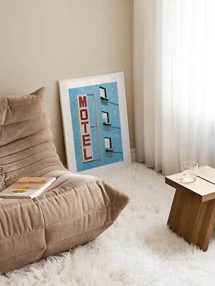 Poster motel incorniciato appoggiato al muro accanto a poltrona beige e tavolino in legno