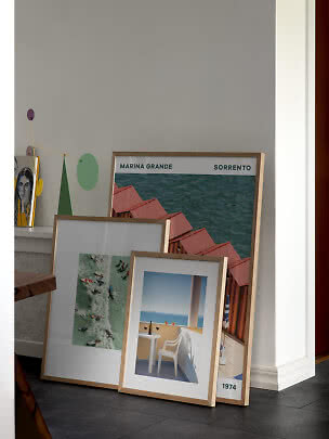 Three framed art prints with light wood frames leaning against a gray wall in a modern home.