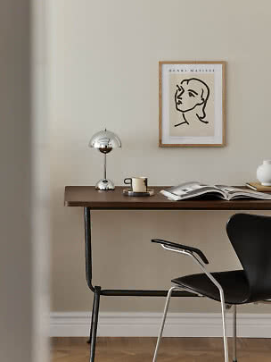 Scrivania con lampada cromata, tazza, libro aperto e sedia nera sotto un poster Matisse incorniciato.