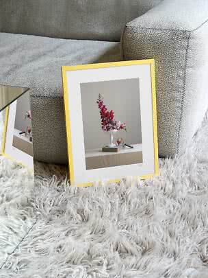 Goldfarbener Bilderrahmen mit Blumenmotiv, an graues Sofa auf weißem Hochflorteppich gelehnt
