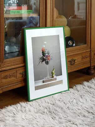 Ingelijste bloemenprint met groene lijst, leunend tegen een houten kast op een wit hoogpolig kleed.