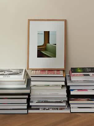 Holzgerahmter Kunstdruck mit grünem Sitz am Fenster, auf Bücherstapeln vor beiger Wand