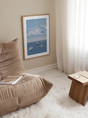 Poster incorniciato con barche a vela sul mare accanto a una poltrona beige e un tavolino in legno