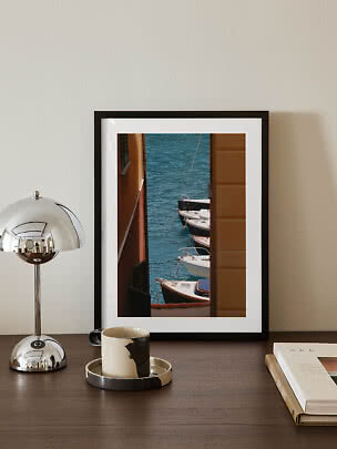 Framed photo of boats by the water on a table with chrome lamp, mug, and book