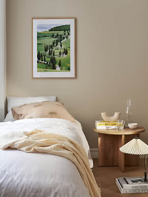 Schlafzimmer mit Bett, rundem Holztisch und gerahmtem Landschaftsbild an beigefarbener Wand