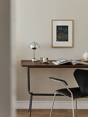 Scrivania in legno scuro con lampada cromata, tazza, libro aperto e sedia nera su parete beige