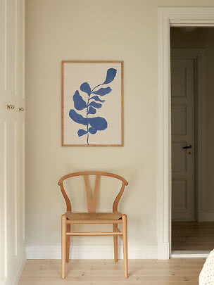 Framed print with blue botanical shape above a light wooden chair on a beige wall