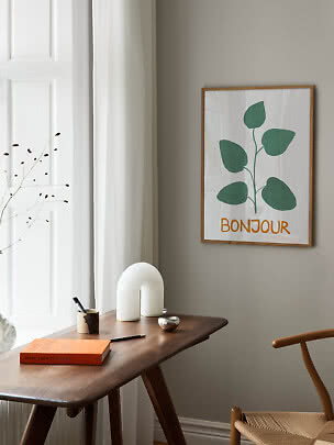 Indrammet plakat med grøn plante og teksten "Bonjour" på grå væg over træbord.