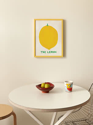 Framed lemon poster on wall above a round white table with fruit bowl and colorful mug