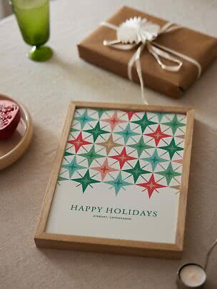 Stampa incorniciata 'Happy Holidays' con stelle colorate su un tavolo accanto a regalo e bicchiere verde