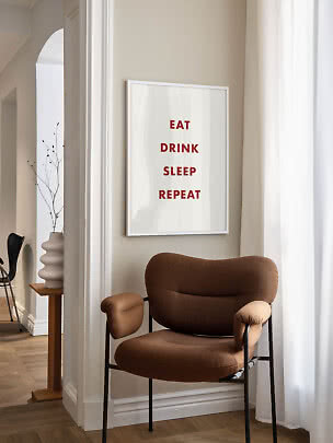 Indrammet plakat med rød tekst over en brun lænestol ved lyse gardiner