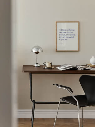 Minimalistisk skrivebord med kromlampe, kop, åben bog og sort stol ved væg med indrammet plakat