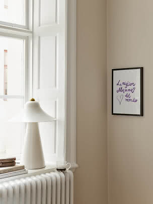 Framed poster with purple text on a beige wall beside a window and white table lamp