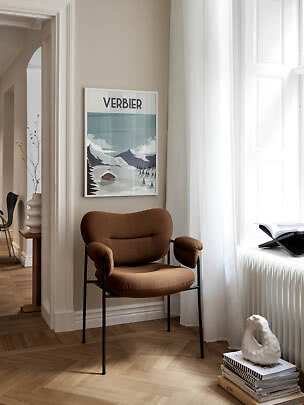 Brun polstret stol med sorte ben ved vindue og Verbier-plakat på beige væg