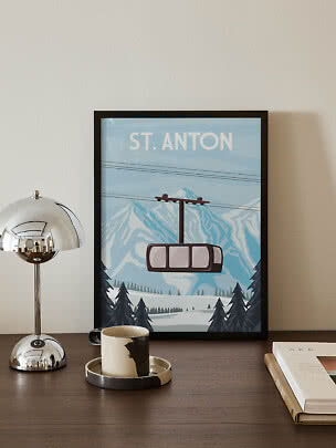 Framed St. Anton mountain poster with cable car on a wooden table beside a lamp