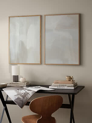To abstrakte plakater i lyse beige toner i trærammer over et skrivebord.