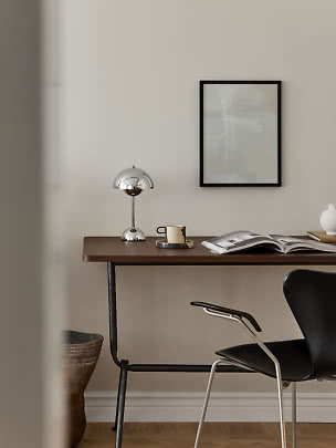 Minimalistisk skrivebord med kromlampe, kop, åben bog og sort stol ved beige væg