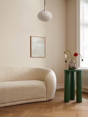 Grønt rundt sidebord med tre ben ved beige sofa, vase med blomster og vindue.