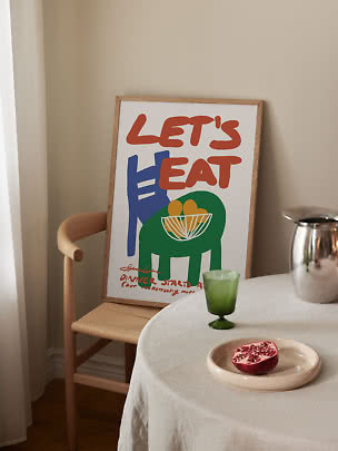 Indrammet farverig plakat med teksten "Let's Eat" lænet mod stol ved dækket bord