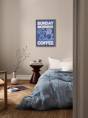 Póster azul enmarcado "Sunday Morning Coffee" sobre una cama en un dormitorio tranquilo.