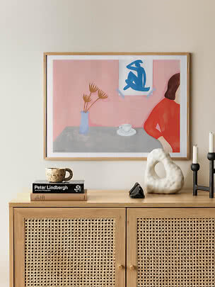 Indrammet abstrakt kunsttryk med rosa baggrund, vase, kop og figur i rødt på egetræsskænk
