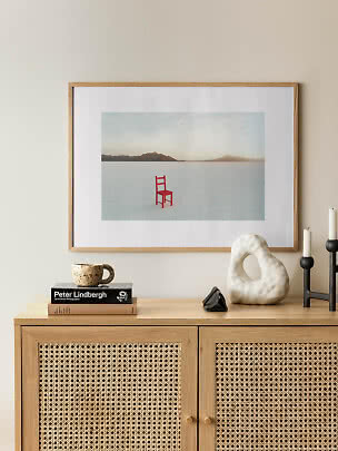 Gerahmter Kunstdruck mit rotem Stuhl in weißer Landschaft über einem Sideboard aus hellem Holz