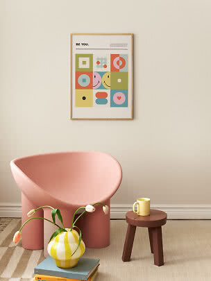 Indrammet farverig plakat med grafiske former og tekst på beige væg over en rosa stol