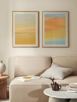Two abstract prints in light wood frames above a beige sofa in a calm living room