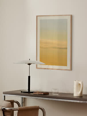 Framed art print with yellow sky above a console table with lamp, glass and cream vase