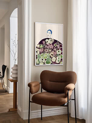 Stampa artistica incorniciata di una donna tra fiori sopra una poltrona marrone in un angolo luminoso