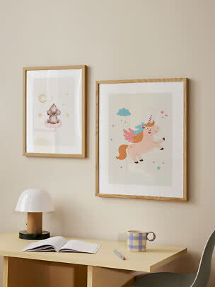 Zwei gerahmte Kinderbilder mit Maus und Einhorn über einem Schreibtisch mit Lampe, Buch und Tasse.