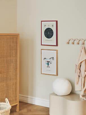 Zwei gerahmte Kinderposter an beigefarbener Wand über einer Bank mit runder weißer Lampe.