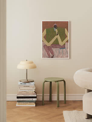 Grøn skammel ved siden af stak bøger med bordlampe under indrammet kunst på beige væg
