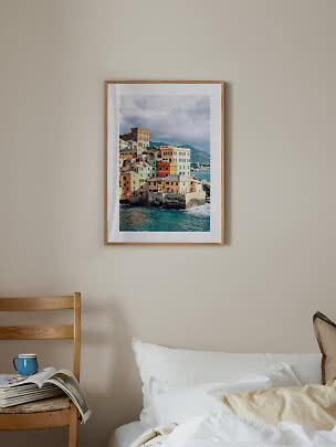 Framed coastal print with colorful houses above the sea on a beige wall over a bed
