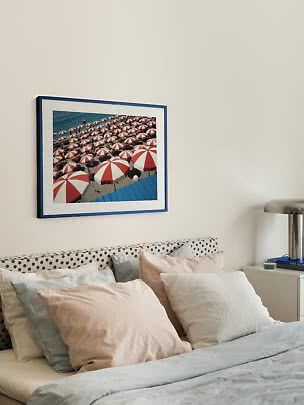 Blue-framed wall art of red and white beach umbrellas above a bed with soft light pillows.