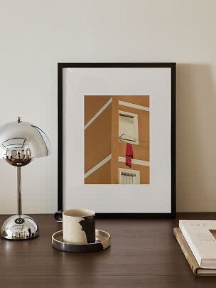 Framed print of an ochre building with a red garment on a clothesline, displayed on a dark table