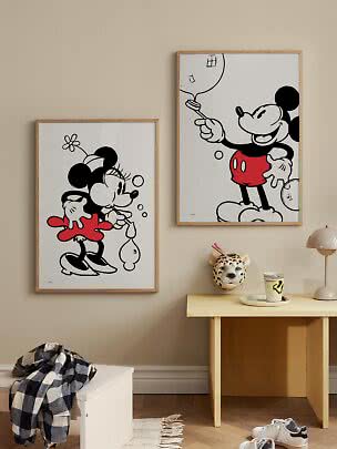 Zwei gerahmte Mickey- und Minnie-Mouse-Poster an einer beigen Wand über einem kleinen Tisch.