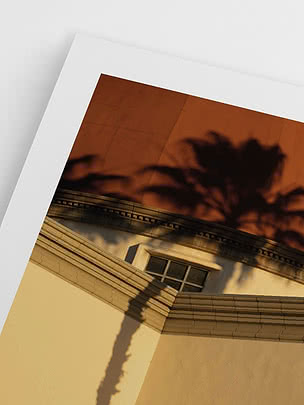 Gerahmtes Foto eines Gebäudes mit gebogenem Gesims, kleinem Fenster und Palmenschatten vor orangem Himmel